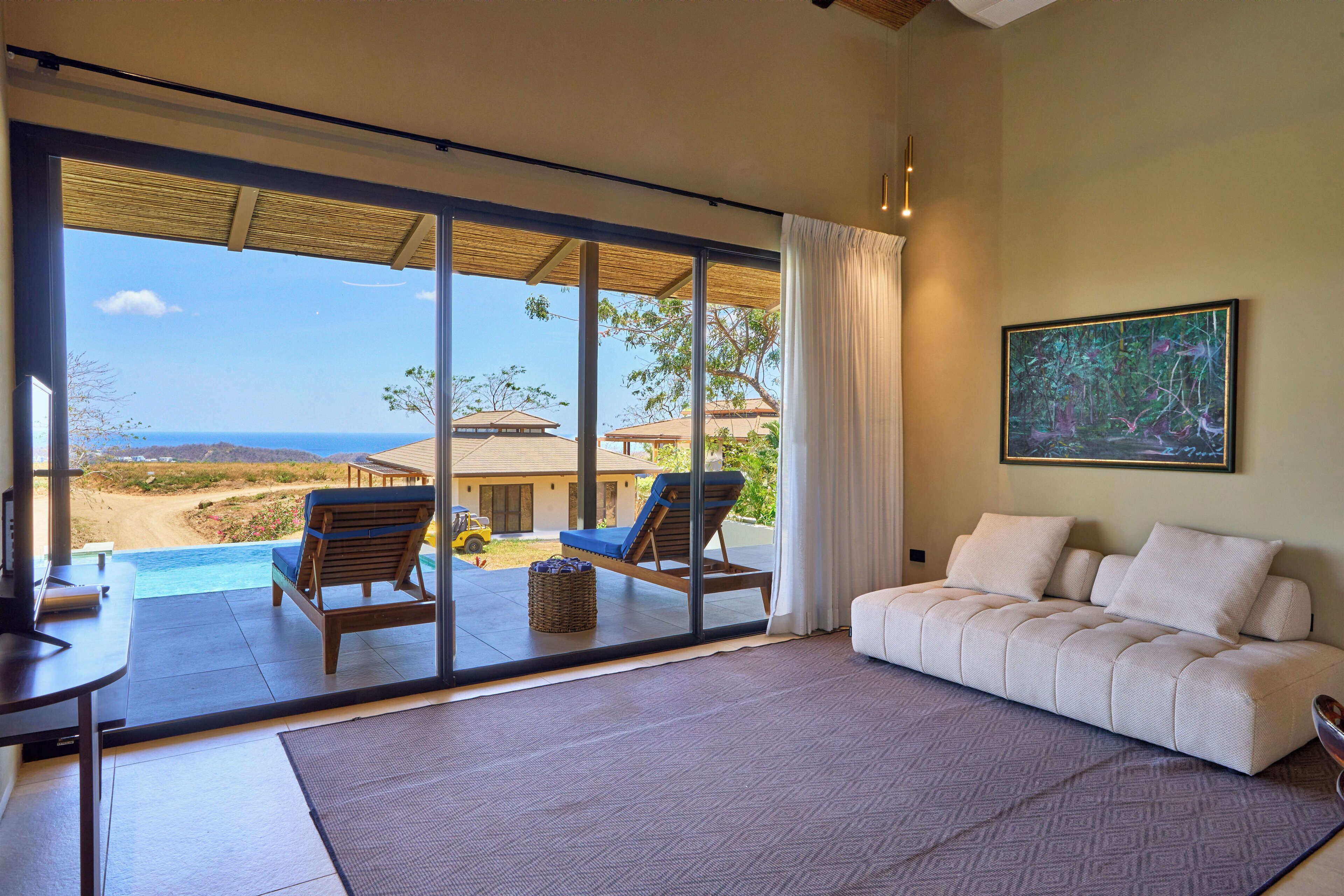 A spacious room with a large sliding glass door leading to a balcony overlooking a beach and ocean. There is a white sofa and a wooden chair on the balcony.
