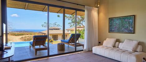 A spacious room with a large sliding glass door leading to a balcony overlooking a beach and ocean. There is a white sofa and a wooden chair on the balcony.