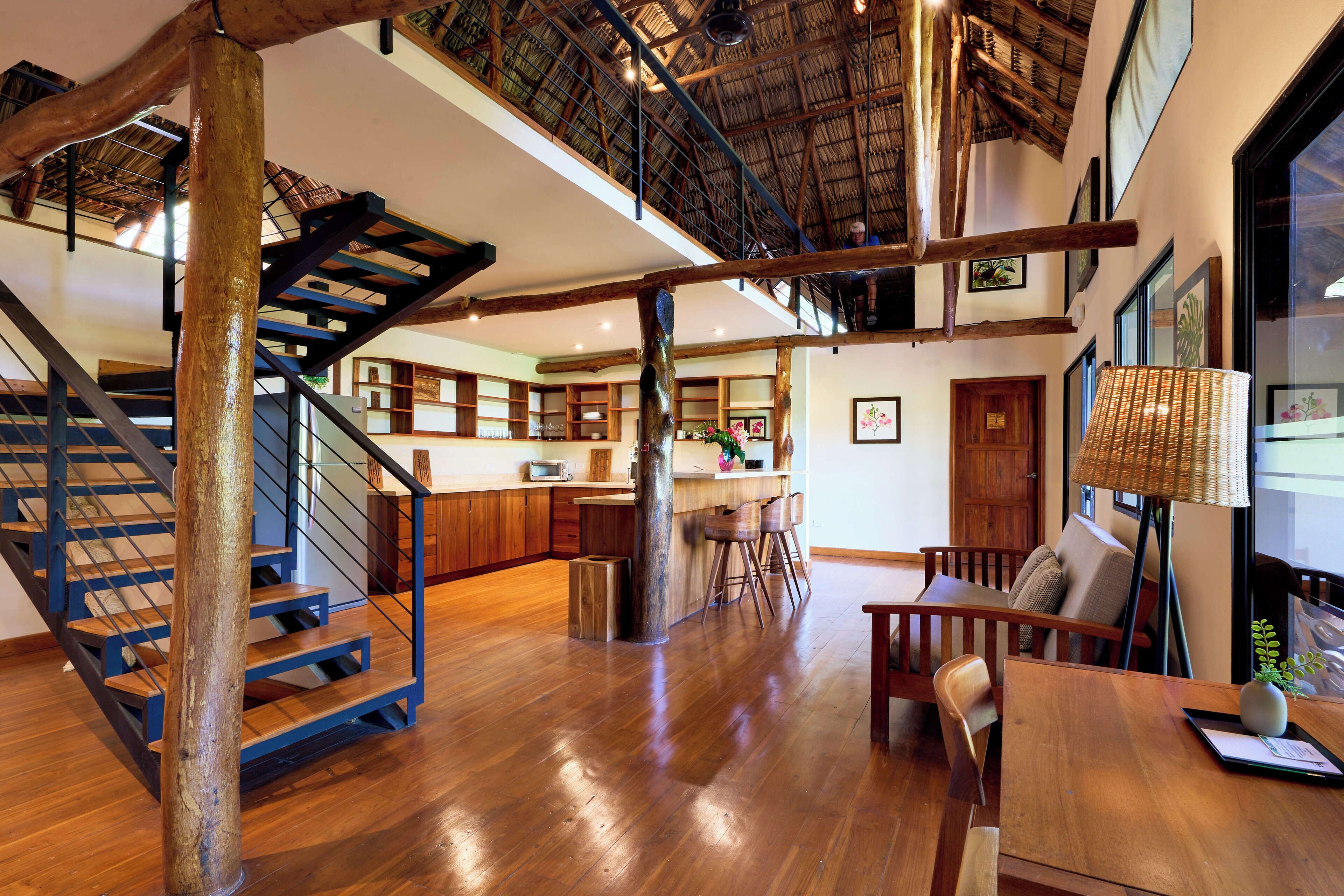 A spacious interior with a wooden staircase, a dining area, and a kitchen with a high ceiling and exposed beams.