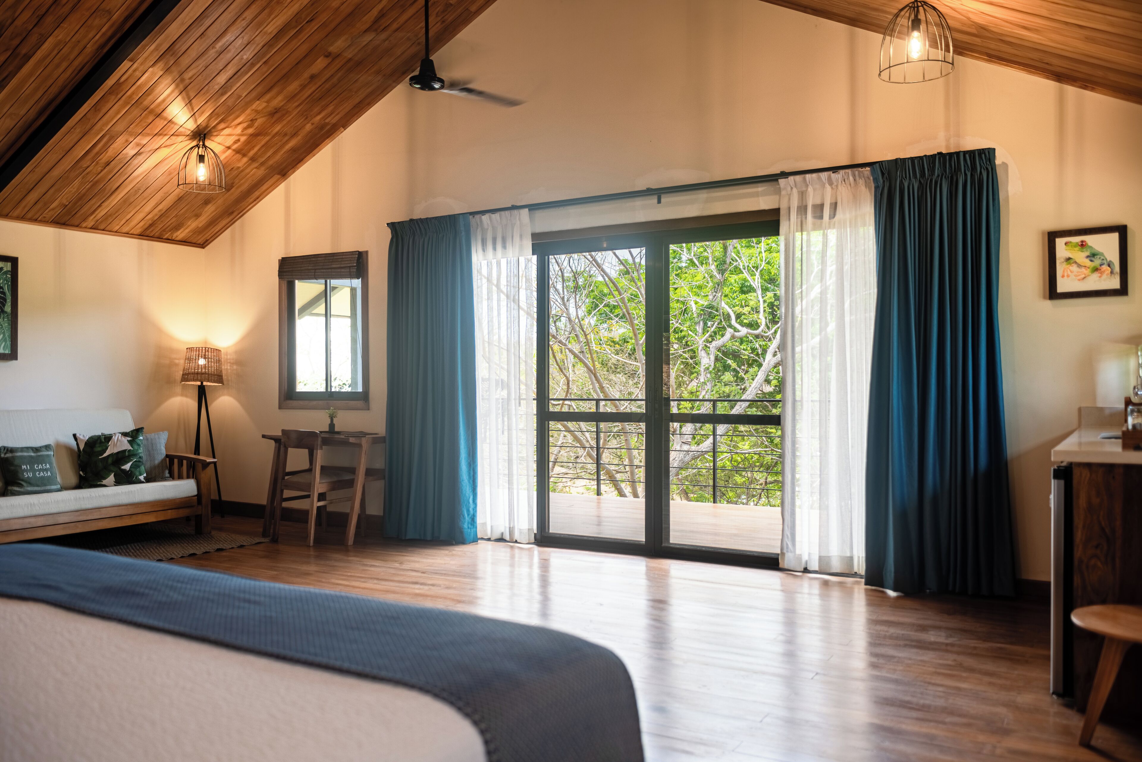 Una habitación amplia con techo de madera, una cama grande, un escritorio y vistas a la vegetación a través de puertas de cristal.