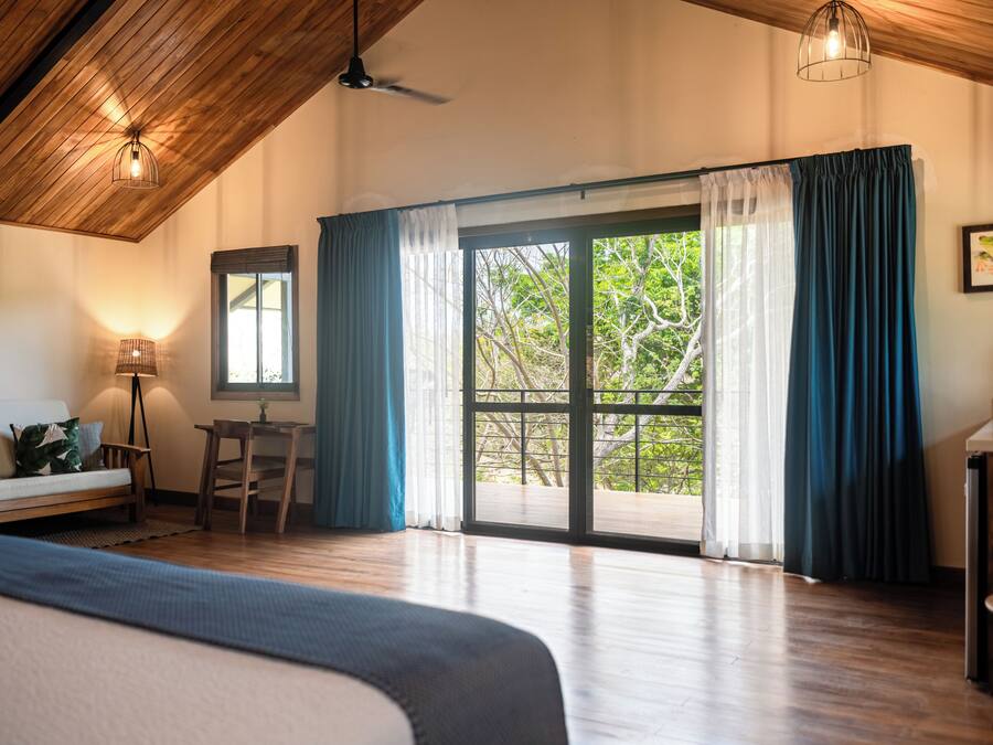Una habitación amplia con techo de madera, una cama grande, un escritorio y vistas a la vegetación a través de puertas de cristal.