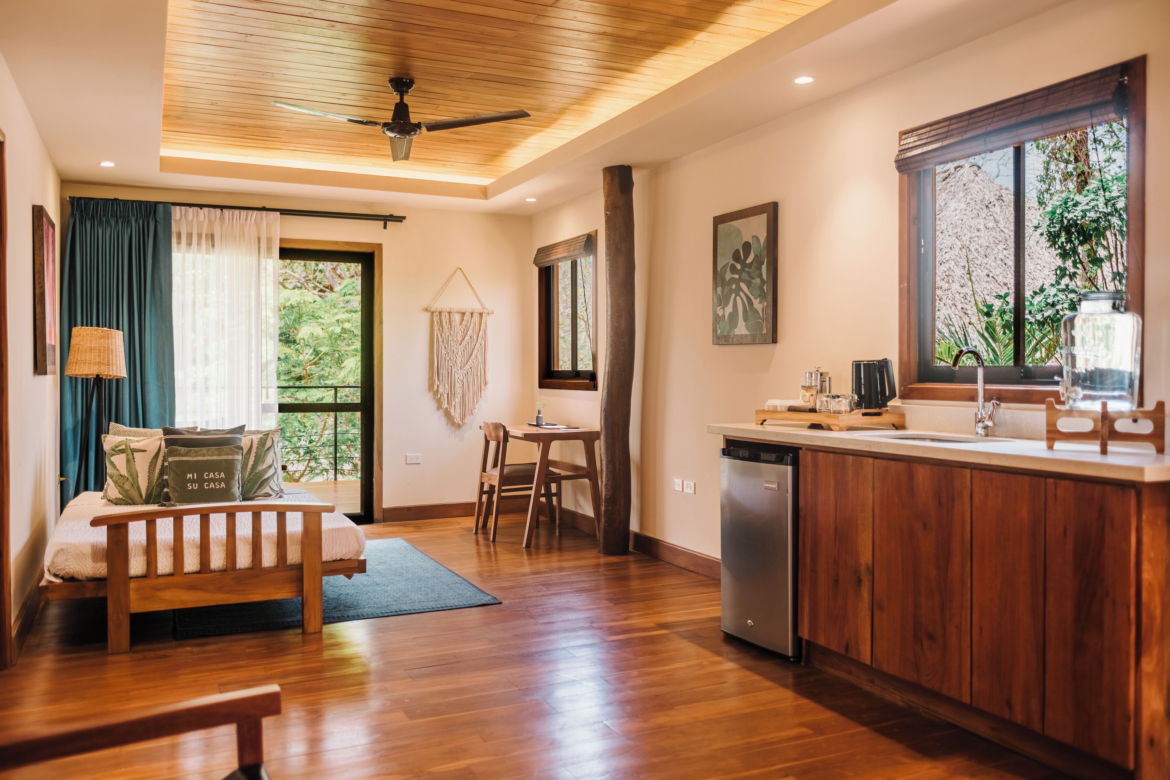 Una habitación moderna con piso de madera, cama con almohadas, escritorio, silla, ventana con cortinas, ventilador de techo y una zona de cocina con pileta y mostrador.