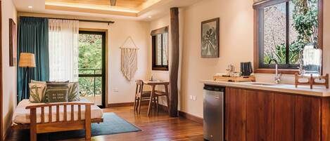 Una habitación moderna con piso de madera, cama con almohadas, escritorio, silla, ventana con cortinas, ventilador de techo y una zona de cocina con pileta y mostrador.