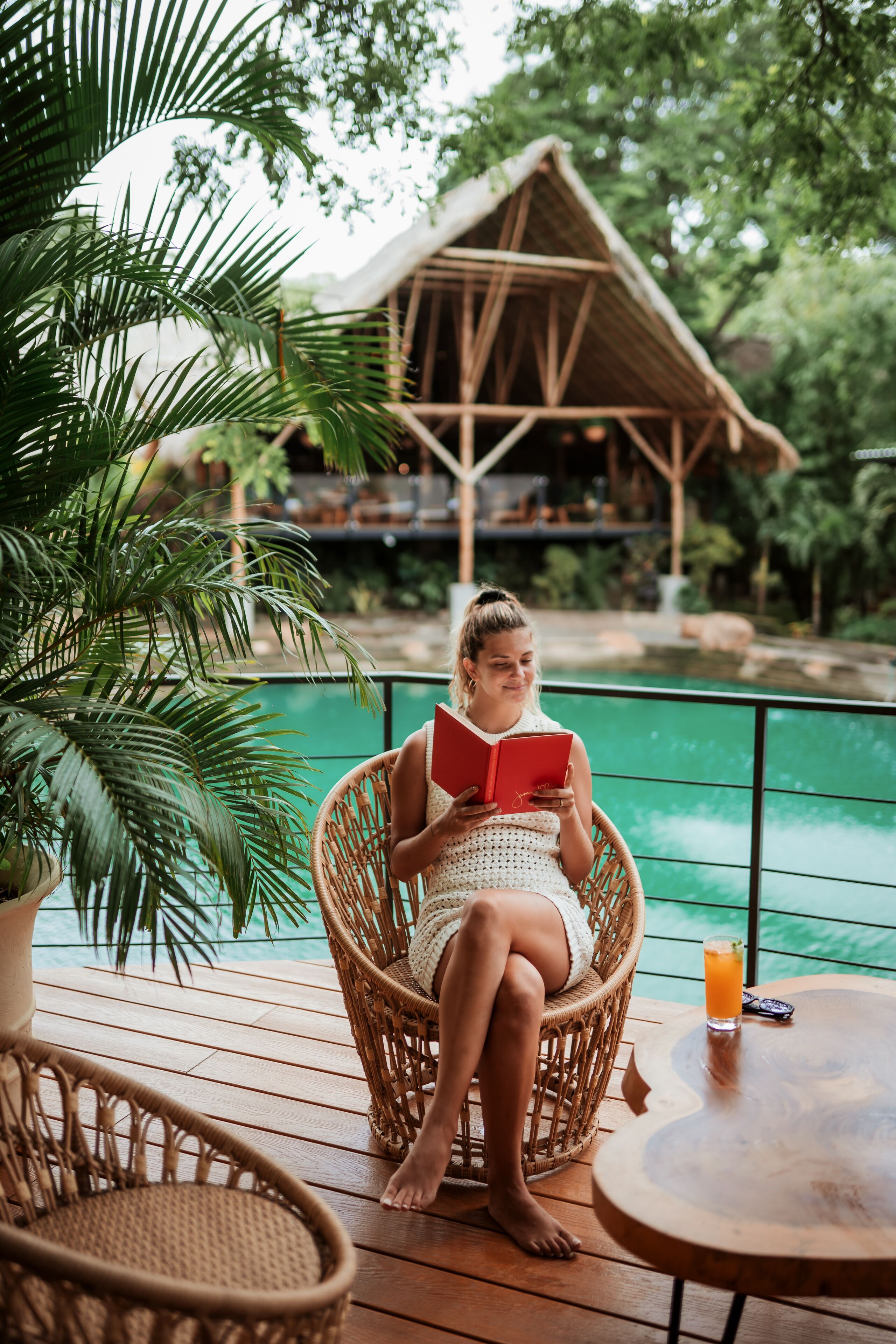Eine Person sitzt auf einem Korbstuhl neben einer Holzterrasse, im Hintergrund ist ein Pool zu sehen.