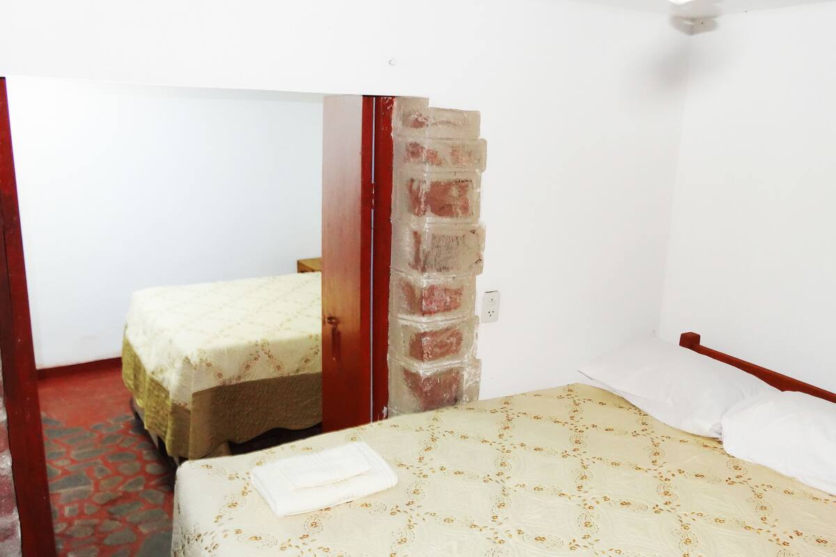 Une petite pièce simple, avec deux lits simples, dont l’un est recouvert d’une couvre-lit à motifs et l’autre d’une couverture unie. On peut voir un mur en briques et un cadre de porte en bois.
