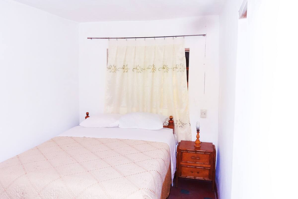 Une petite chambre simple, avec un lit simple, une table de chevet et un rideau.