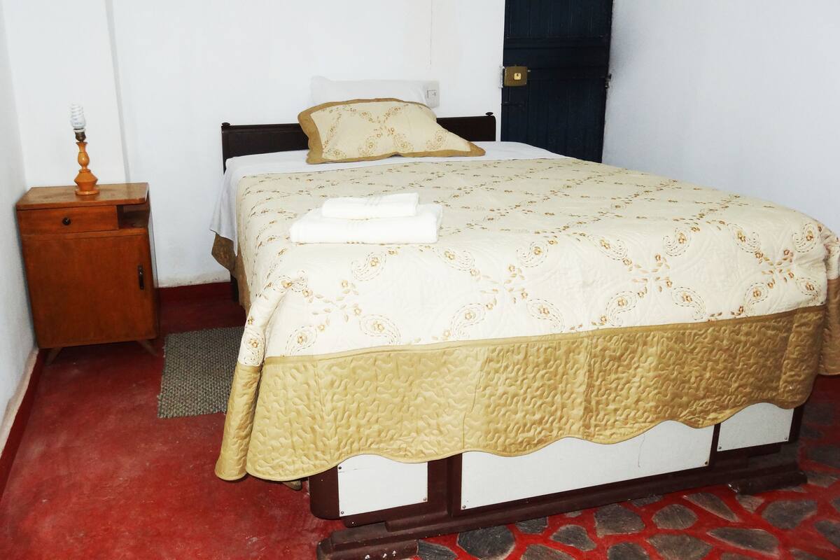 Un lit simple avec une couvre-lit à motifs floraux et un oreiller assorti, une table de chevet en bois avec une lampe, et un sol à motifs rouges et gris.