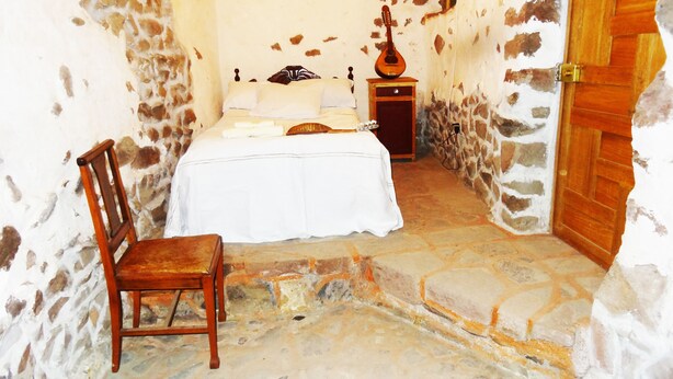 Una habitación estrecha con una cama, una silla, una guitarra y una puerta de madera.