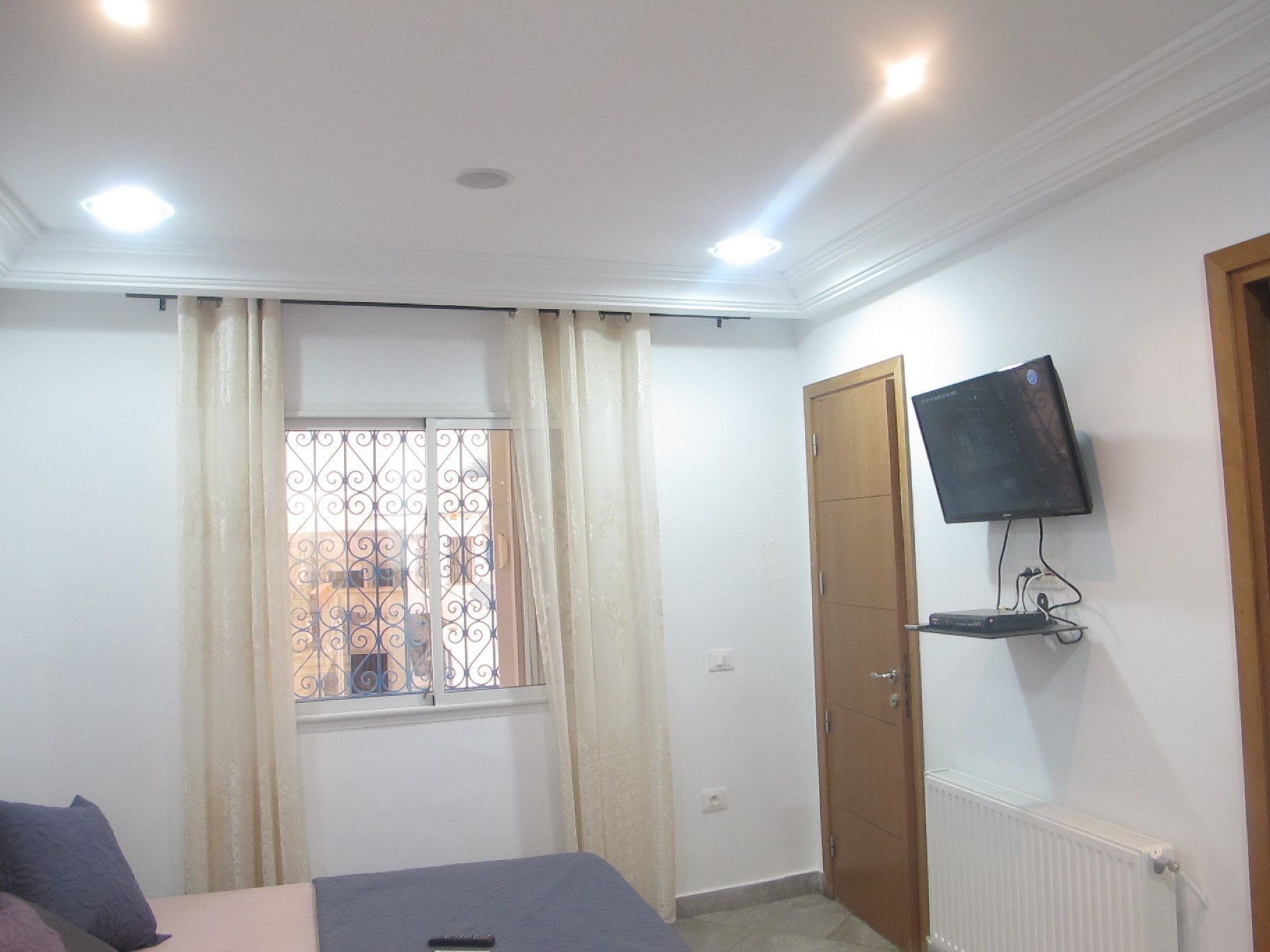A bedroom with a bed, a TV mounted on the wall, a window with curtains, and a radiator.