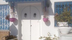 Una puerta blanca con toldo azul, dos plantas en macetas a cada lado y un portón de metal decorativo.