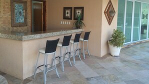 A bar area with a counter and four bar stools, a potted plant, and framed pictures on the wall. - Palm Beach Aquiraz (Aquiraz)
