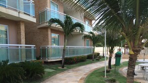 A courtyard with a paved walkway, palm trees, and a building with a glass balcony. - Palm Beach Aquiraz (Aquiraz)