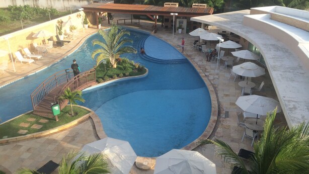 A swimming pool with a curved design, surrounded by lounge chairs and umbrellas, and a covered seating area with tables and chairs. - Palm Beach Aquiraz (Aquiraz)