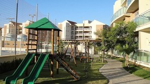 A playground with slides and swings, surrounded by a paved walkway and greenery, with residential buildings in the background. - Palm Beach Aquiraz (Aquiraz)