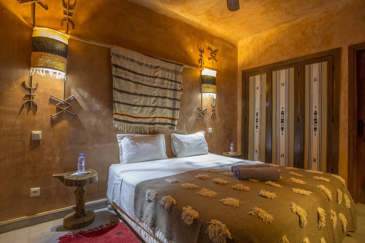 A bedroom with a bed, pillows, a blanket, a nightstand, a water bottle, and wall-mounted lights.