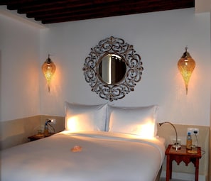 A hotel room with a large bed, two wall-mounted lamps, a decorative mirror, and a bedside table with a bottle and glass.