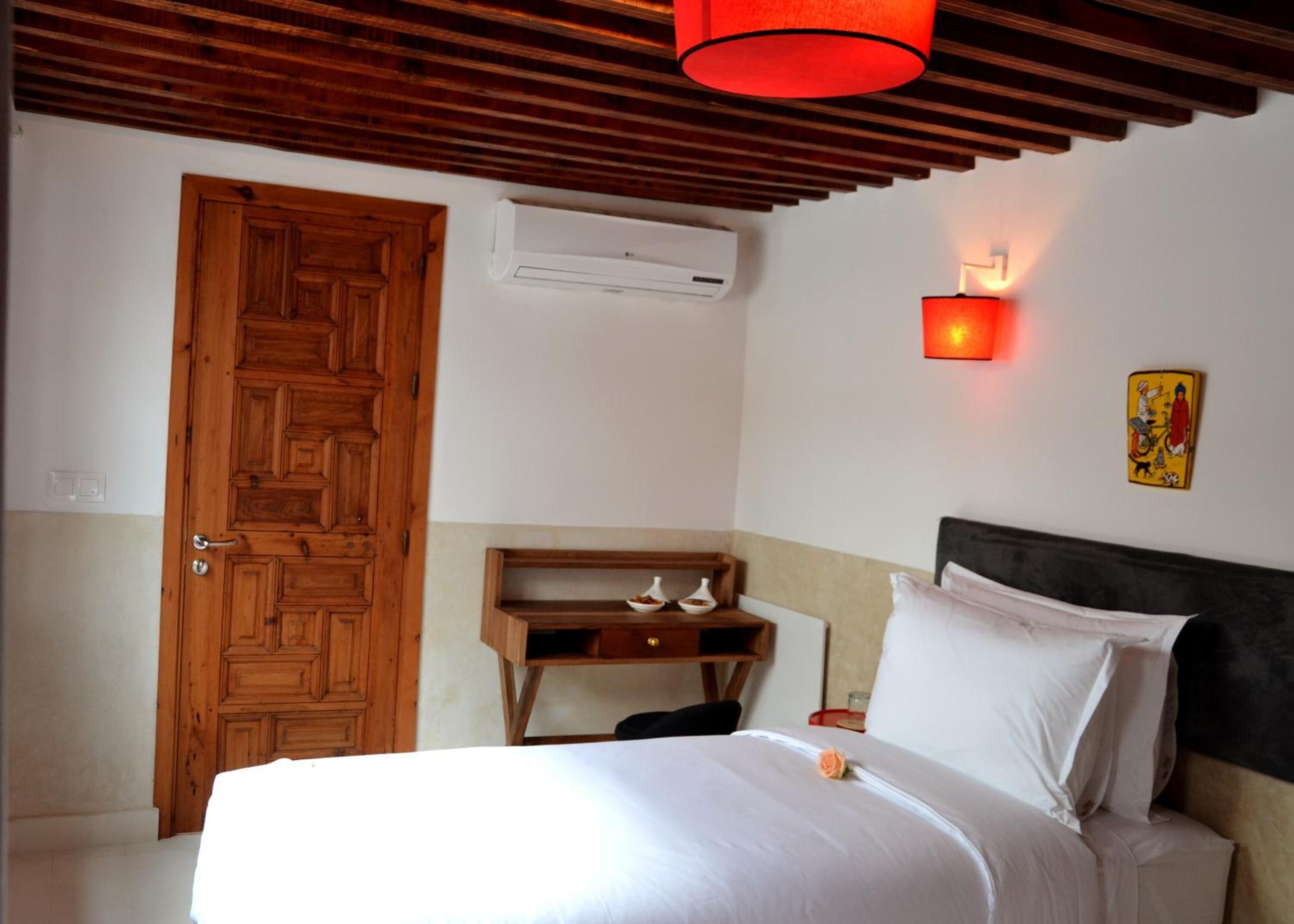Un dormitorio con puerta de madera, un equipo de aire acondicionado, una mesita de noche con lámpara y una cama con ropa de cama blanca.
