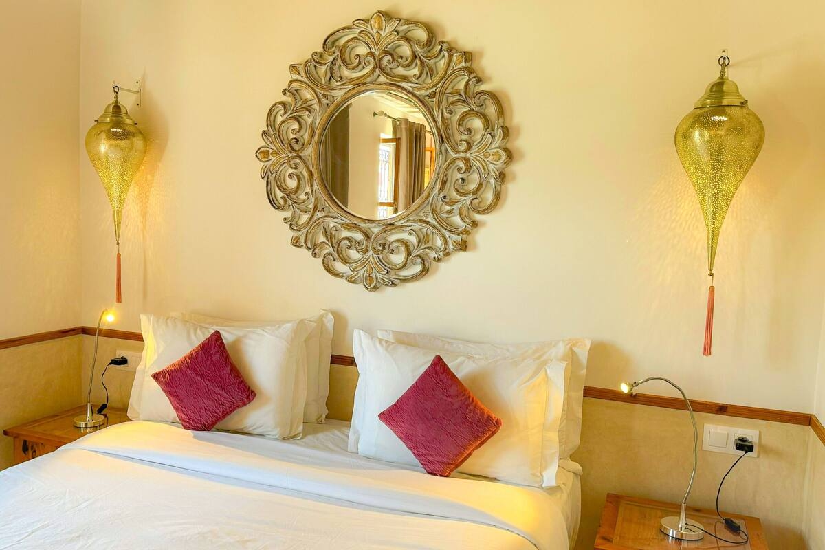 A hotel room with a large ornate mirror, two wall-mounted lamps, a bed with white linens and two red pillows, and bedside tables with lamps and power outlets.