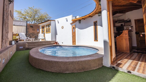 Une piscine entourée d’un petit jardin avec du gazon synthétique, une terrasse en bois et un bâtiment à l’extérieur blanc, agrémenté de détails en bois.