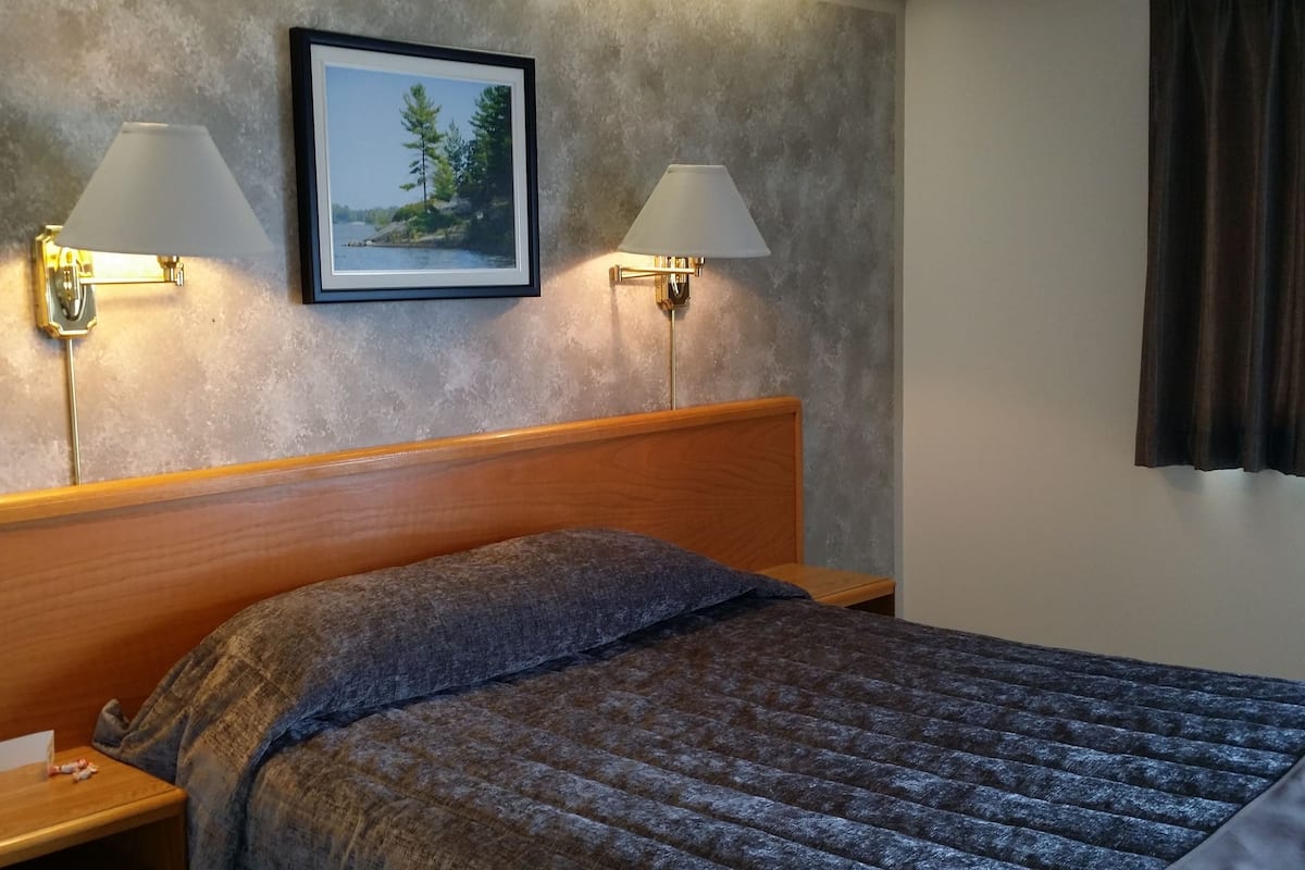 Ein Hotelzimmer mit hölzernem Kopfteil, einem Bett mit grauer Decke, zwei Nachttischlampen, einem gerahmten Bild an der Wand und einem Nachttisch mit Telefon und einem kleinen Gegenstand.