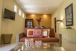 Uma sala de estar com sofá, dois vasos decorativos, uma televisão de tela plana, uma cozinha ao fundo e um quadro na parede.