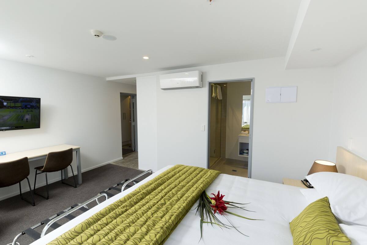 A hotel room with a bed, a desk, a chair, an air conditioning unit, and a television.