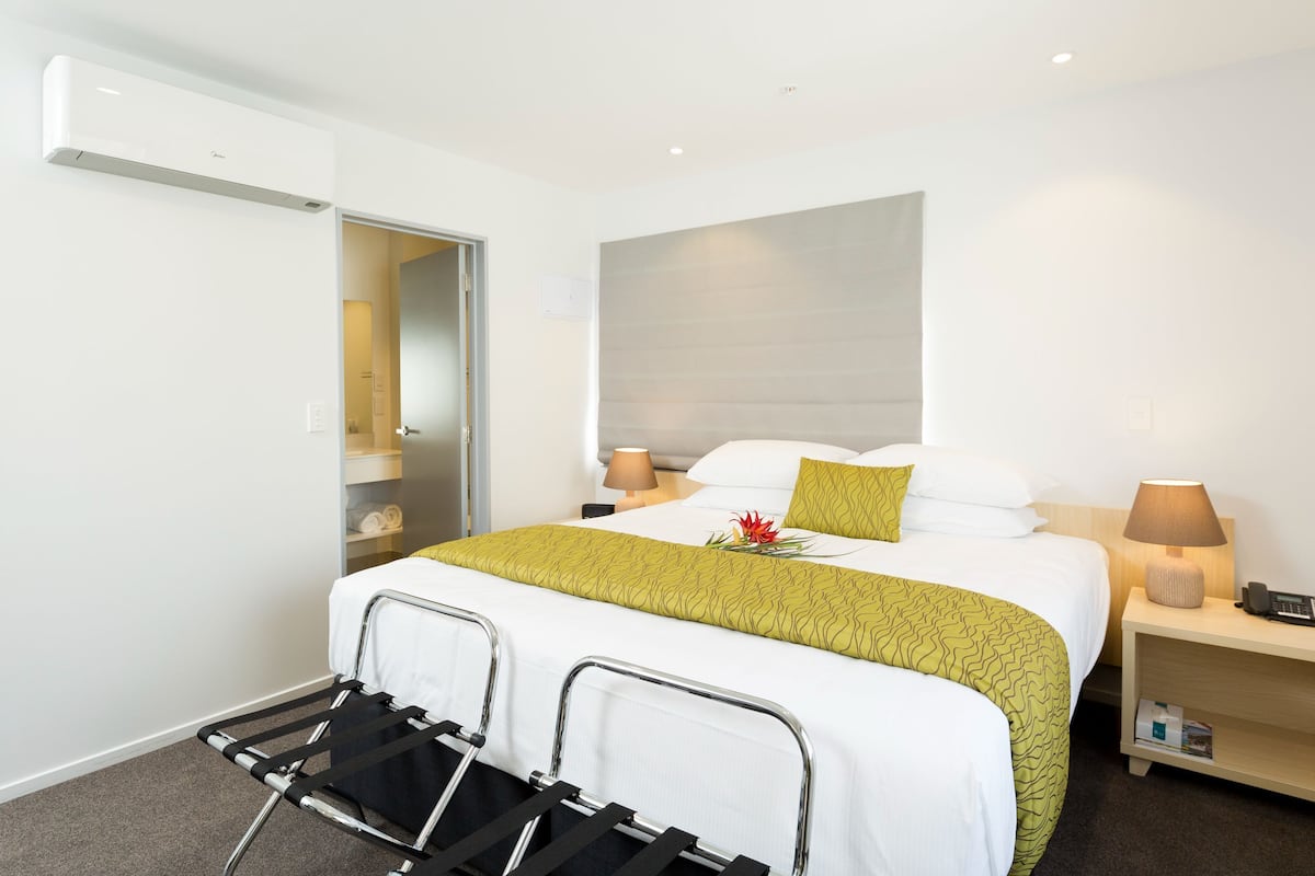 A hotel room with a bed, bedside tables, a lamp, a chair, and an air conditioning unit.