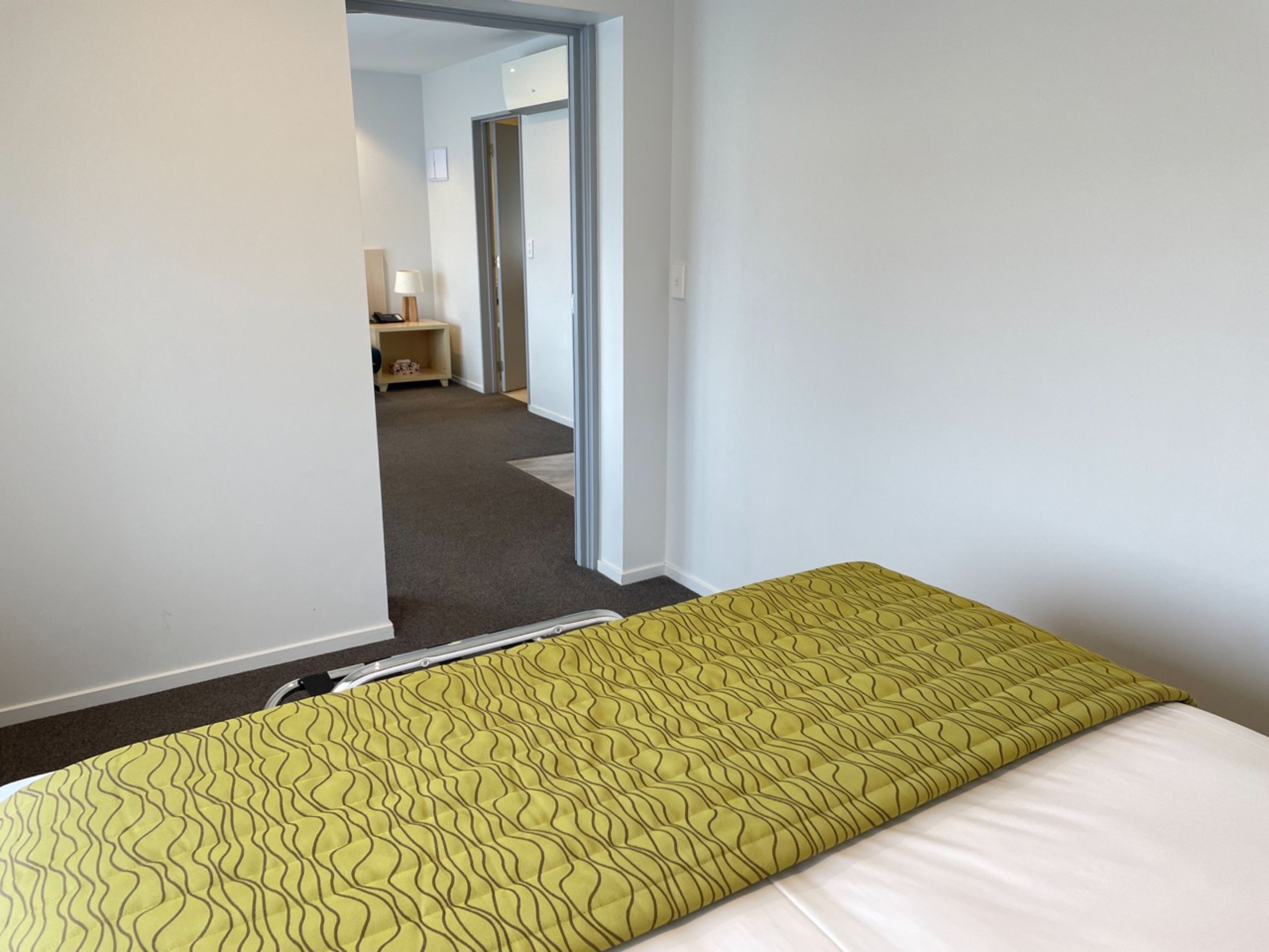 Habitación de hotel con una cama, una mesita de noche, una lámpara y vistas al pasillo.