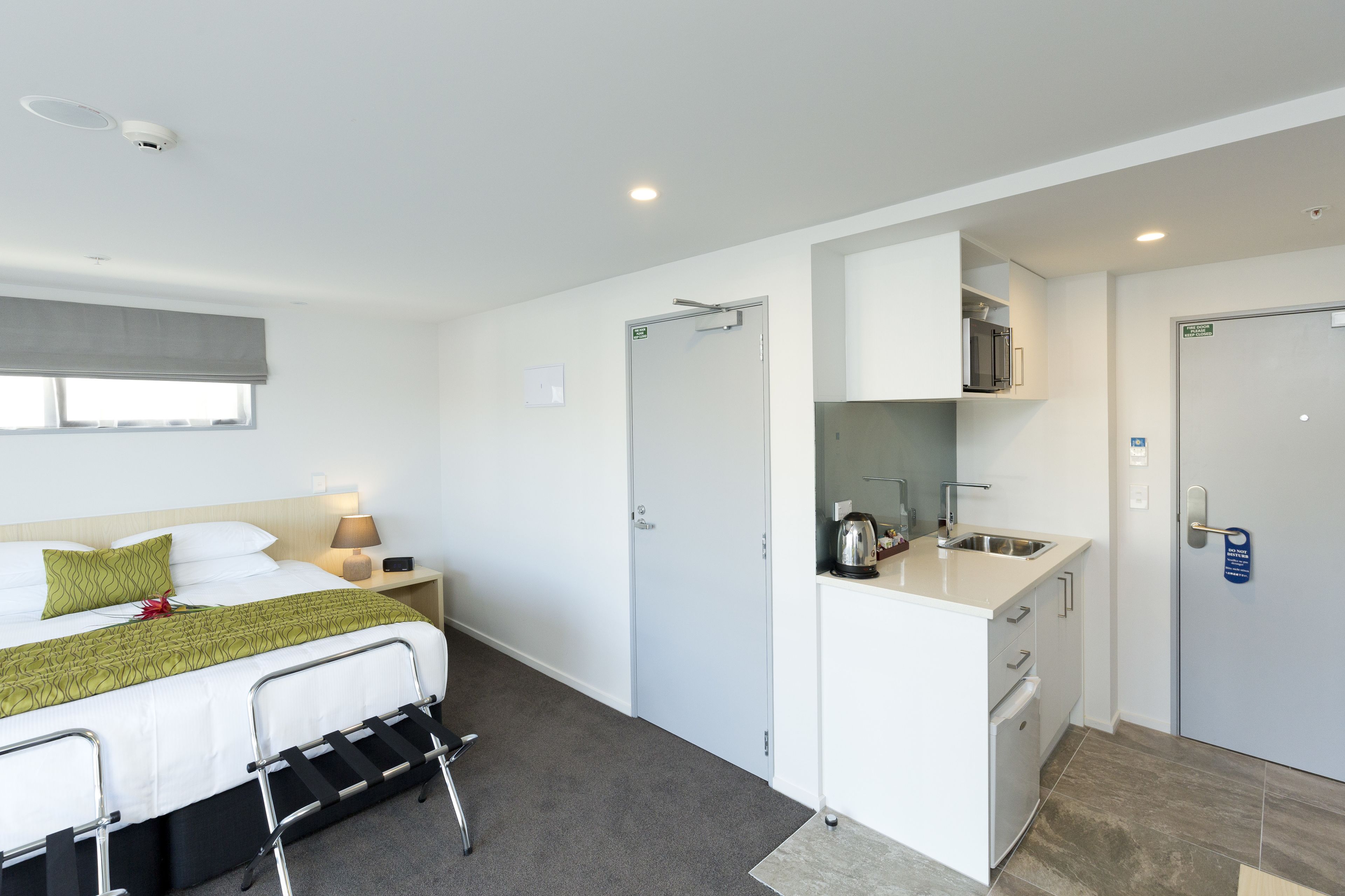 A hotel room with a bed, bedside table, lamp, and a kitchenette area including a sink and cabinets.