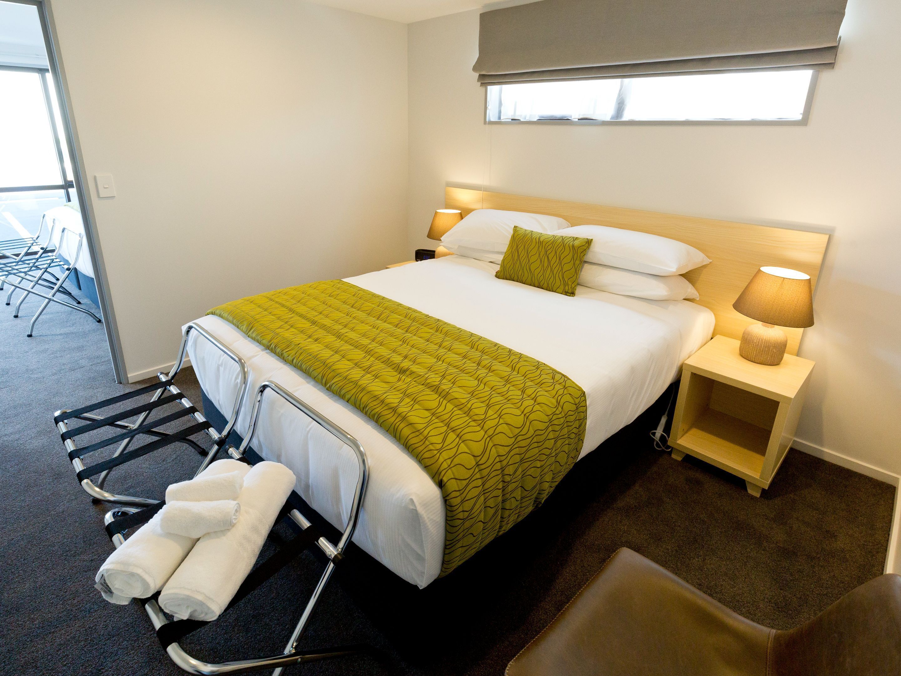 Una habitación de hotel con cama, mesita de noche con lámpara, una silla y una pequeña mesa con toallas.