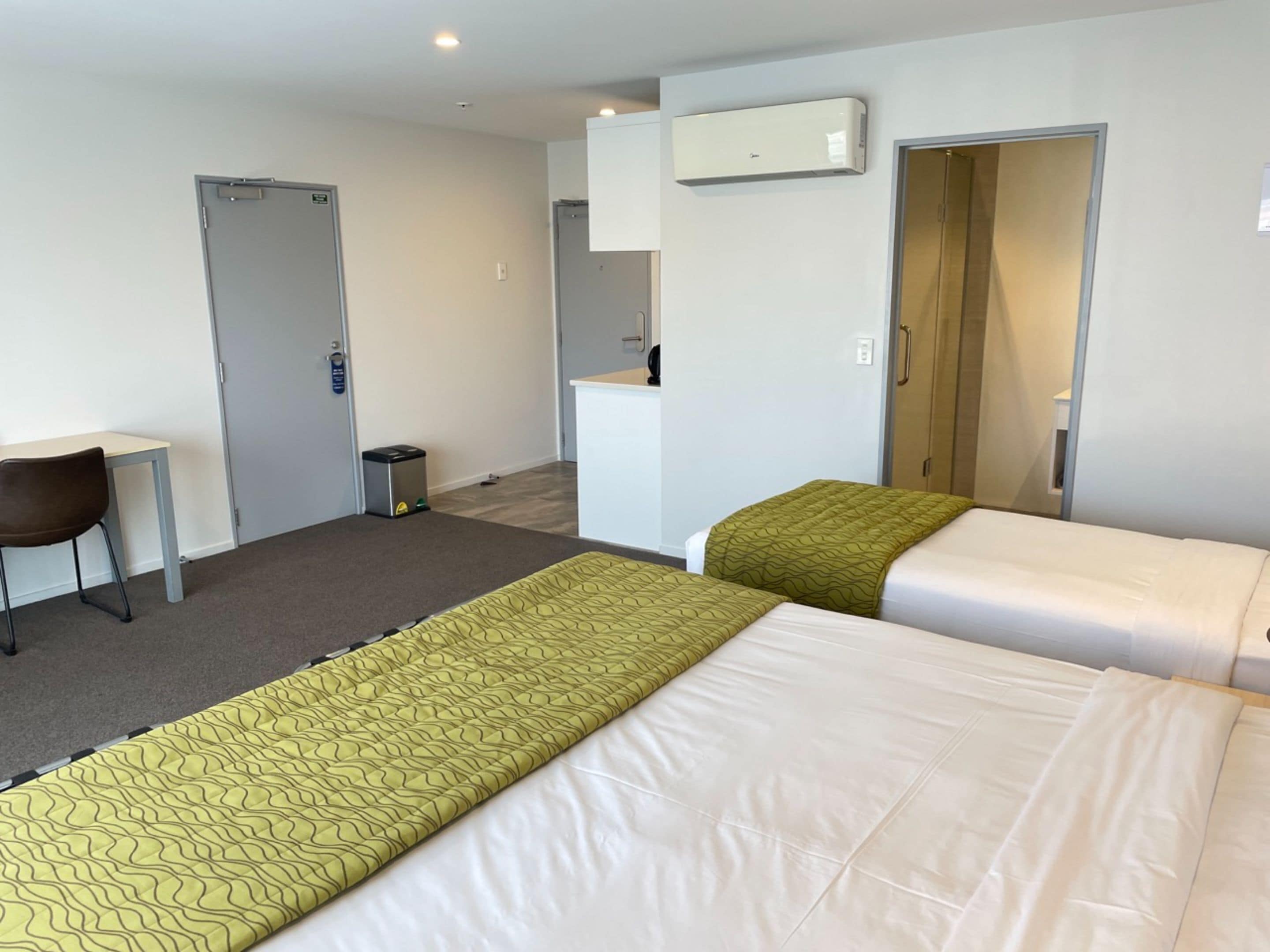 A hotel room with two beds, a desk, a chair, a trash bin, and an air conditioning unit.