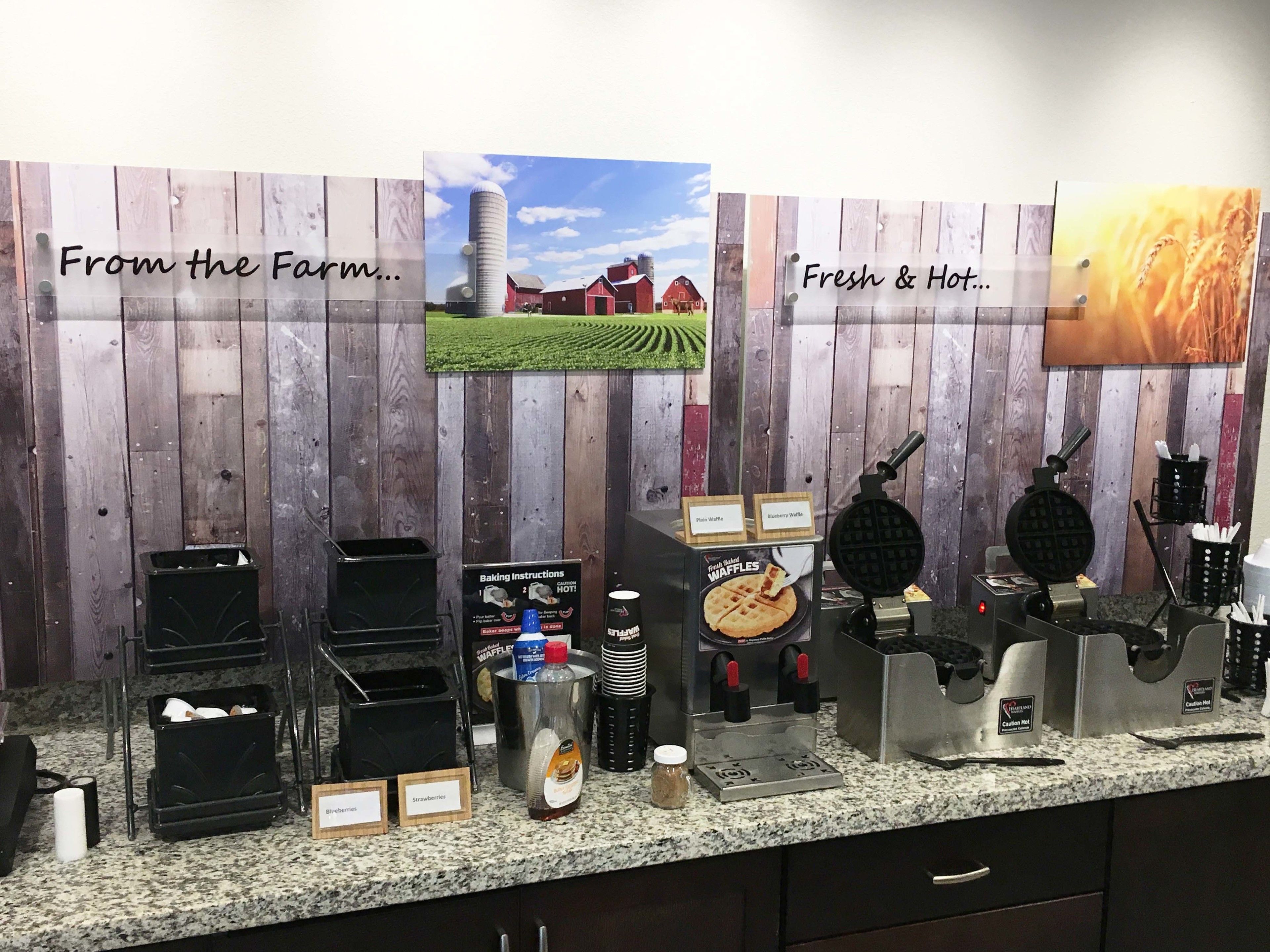 A kitchen counter with waffle makers, waffle batter, and waffle plates. There are signs with text and images on the wall behind the counter.