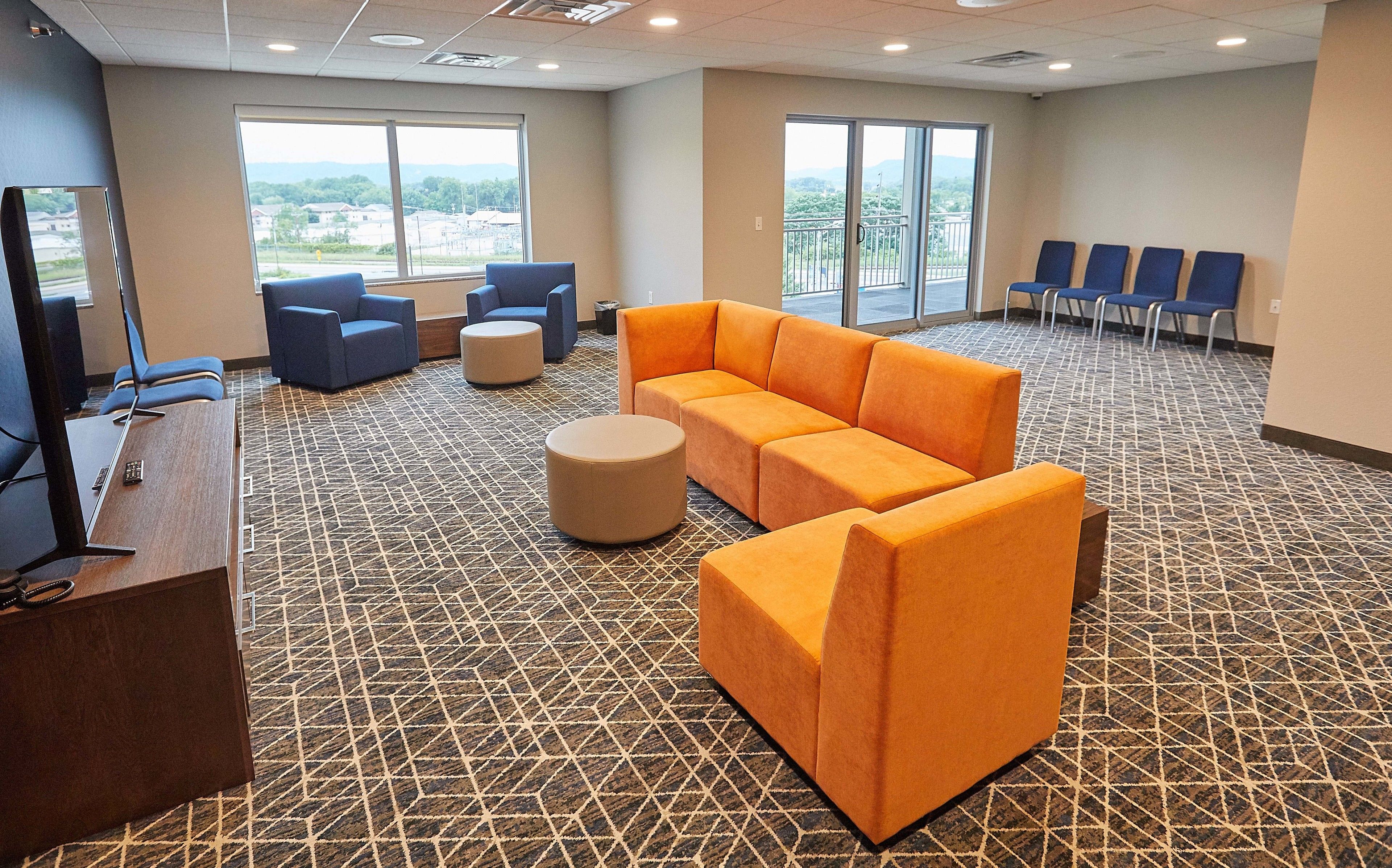 A hotel lobby with a patterned carpet, a reception desk, a large orange sofa, blue chairs, and a view through the windows.