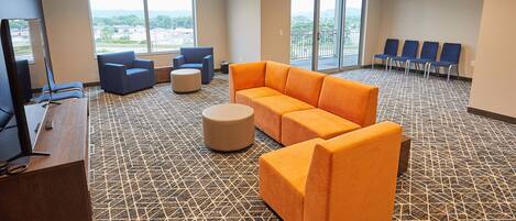 A hotel lobby with a patterned carpet, a reception desk, a large orange sofa, blue chairs, and a view through the windows.