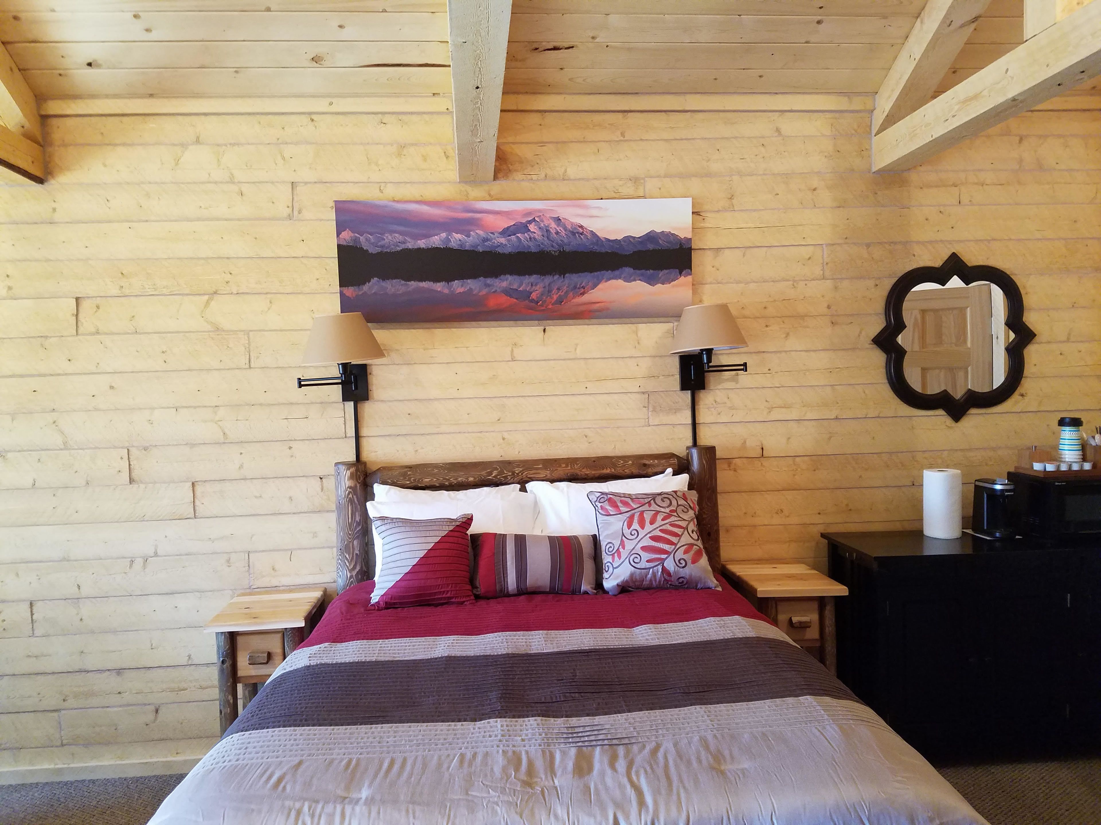 A wooden-paneled room with a bed, two bedside lamps, a painting of a mountain landscape, and a mirror on the wall.