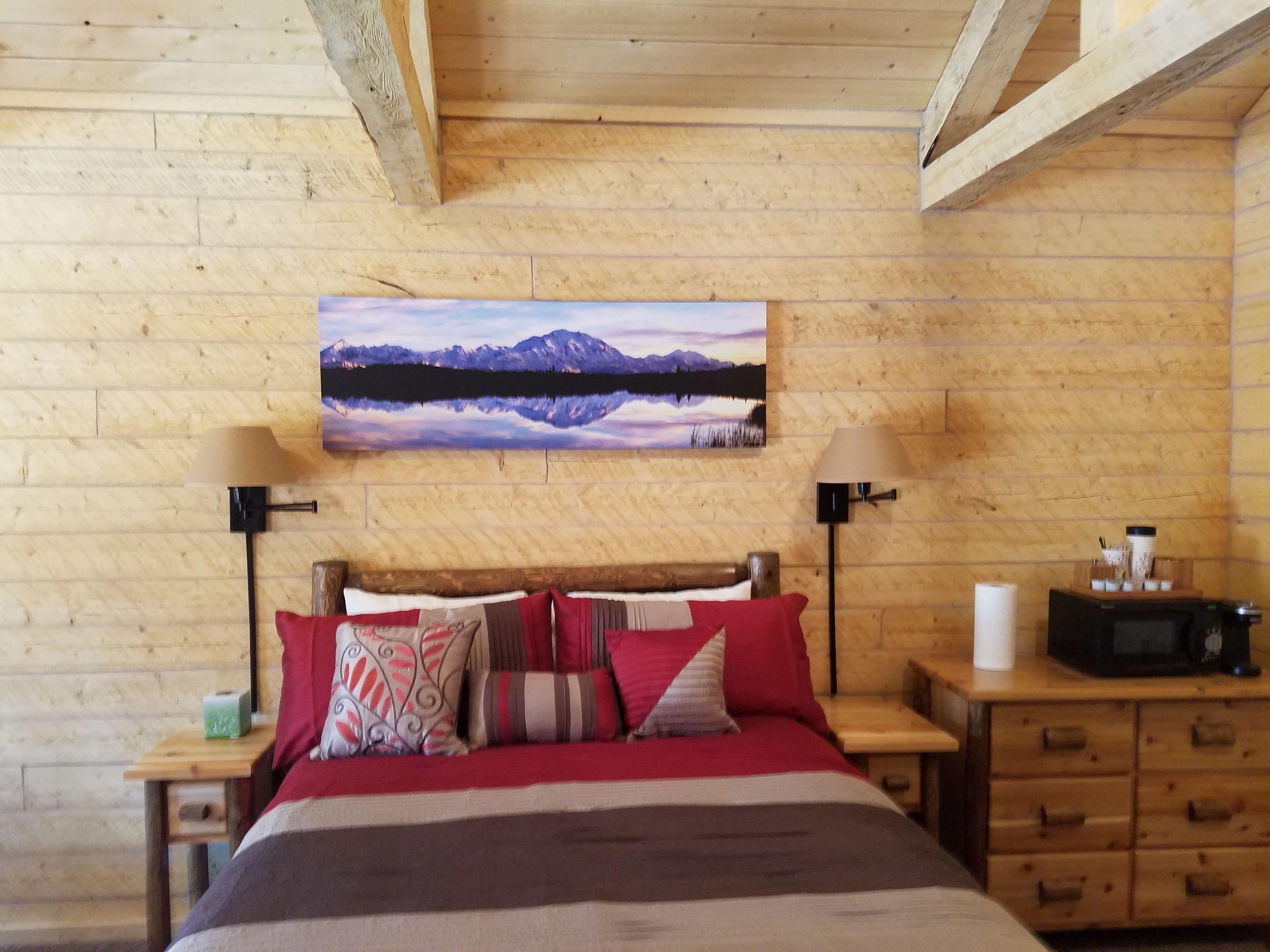 A wooden-paneled bedroom with a bed, two bedside tables, a nightstand, a lamp, a painting, and a microwave.