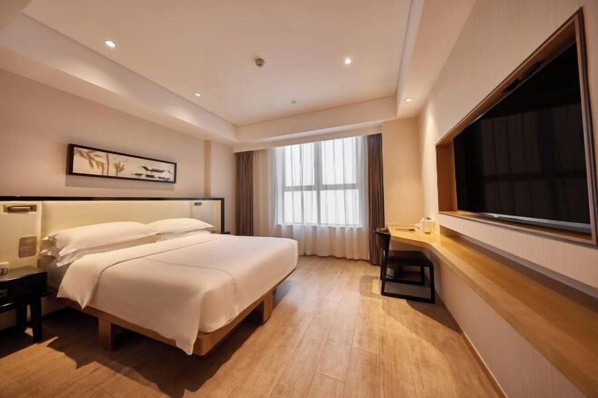 A hotel room with a large bed, a desk, a chair, a window, and a painting on the wall.