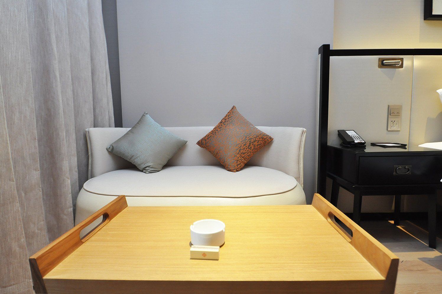 A hotel room with a wooden table, a white sofa with two patterned cushions, a black nightstand with a phone, and a mirror on the wall.