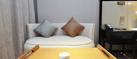 Habitación de hotel con una mesa de madera, un sofá blanco con dos cojines estampados, una mesita de noche negra con un teléfono y un espejo en la pared.