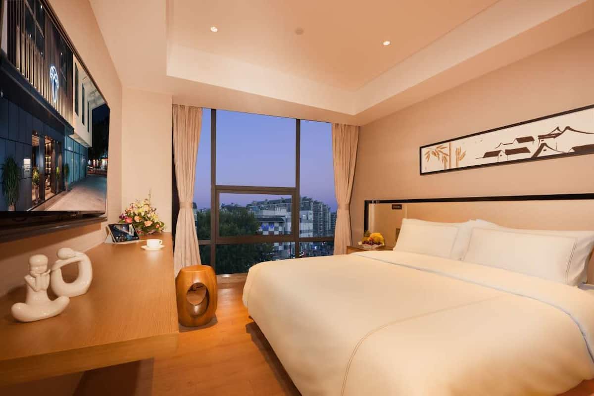 Quarto de hotel moderno com uma cama grande, vista para a cidade através de amplas janelas, televisão de tela plana e mesa de cabeceira de madeira com objetos decorativos.