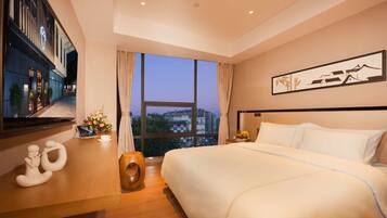 A modern hotel room with a large bed, a view of the cityscape through large windows, a flat-screen TV, and a wooden side table with decorative items.