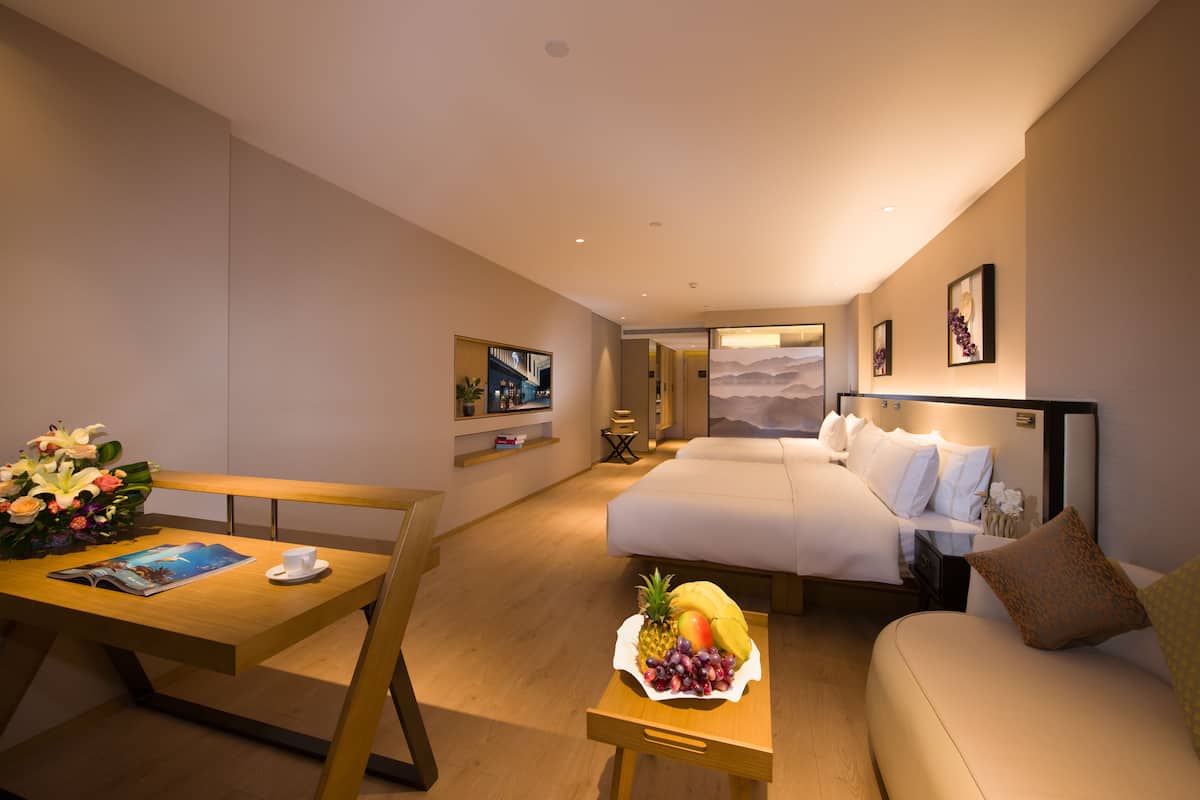 Quarto de hotel moderno com cama, sofá, mesa de jantar com cesta de frutas e televisão na parede.