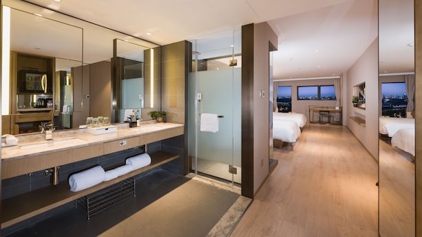 Ein modernes Hotelzimmer mit einem großen Spiegel, einem Schminktisch mit Waschbecken, einer Dusche, einem Bett und einem Schreibtisch mit Blick auf die Stadt.