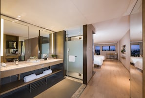 Habitación de hotel moderna con un espejo grande, un tocador con lavamanos, ducha, cama y escritorio con vista a la ciudad.