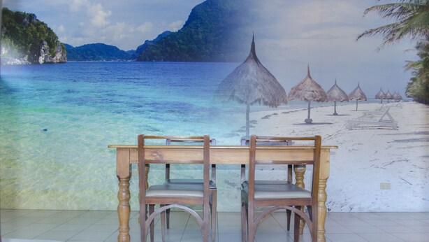 A beach scene with a wooden table and chairs in the foreground, a clear blue sea, and a tropical landscape with mountains and thatched umbrellas in the background. - Tarasa Hostel (Pasay)