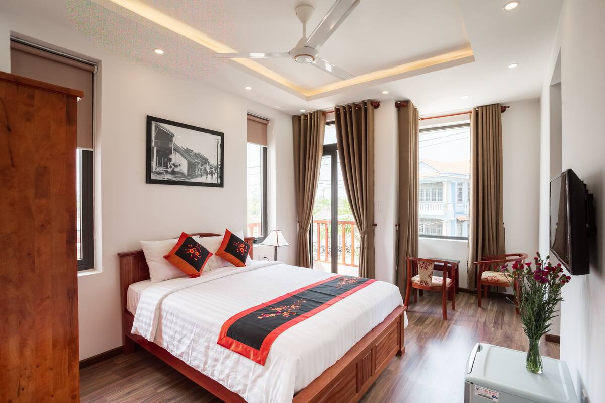 A hotel room with a large bed, wooden headboard, a flat-screen TV mounted on the wall, a small table with chairs, and a window with curtains.