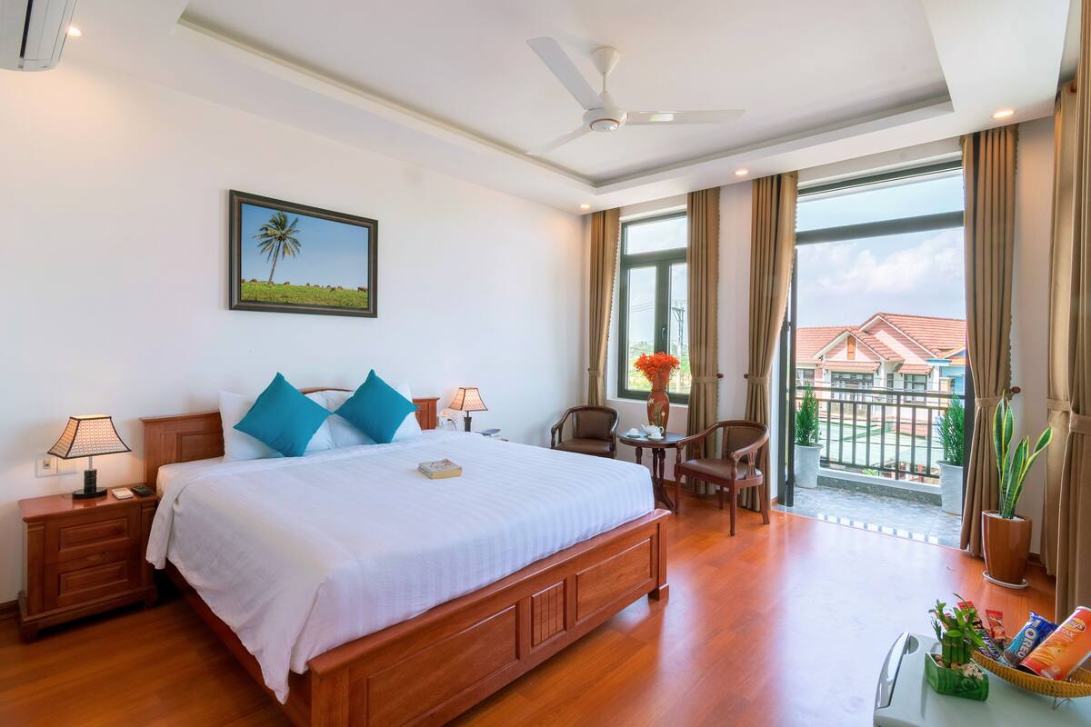 A hotel room with a large bed, a ceiling fan, a view of the outside, and a balcony with plants.