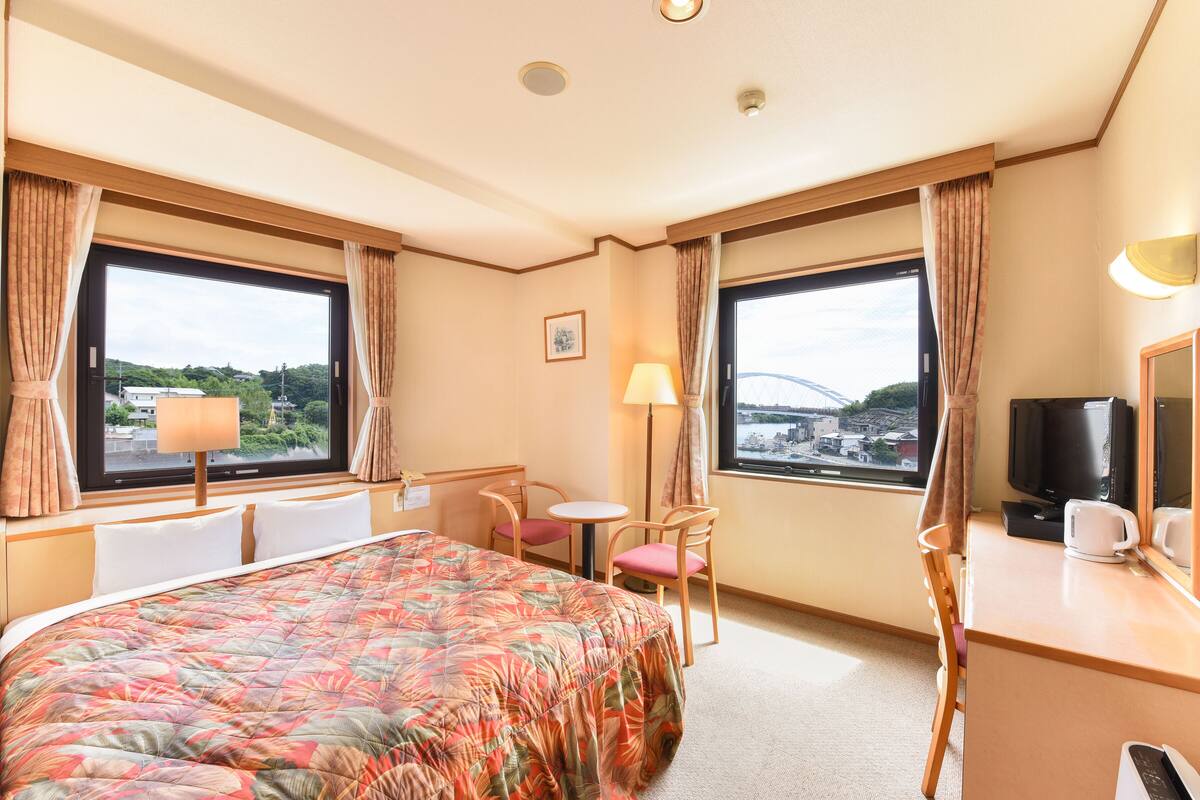 A hotel room with a bed, a desk, a chair, a TV, and large windows offering a view of the city.