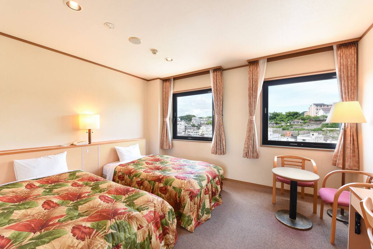 A hotel room with two beds, a small table, a chair, a TV, and a window with a view of buildings and greenery.