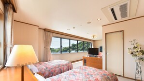 Habitación de hotel con dos camas, un escritorio con lámpara, cómoda, televisión y un amplio ventanal con vistas a zonas verdes.